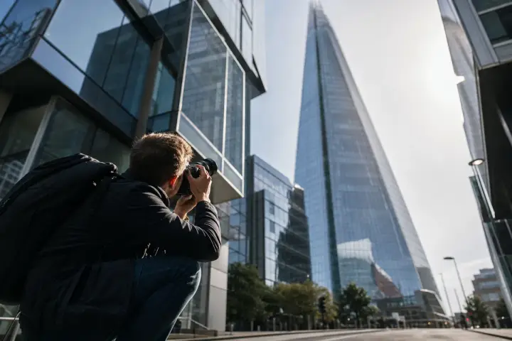 Fotógrafo agachado na rua aponta a câmera para cima, na direção de um arranha-céu de vidro, em ângulo baixo
