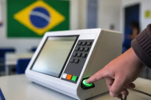 Dedo indicador no leitor de digital de uma urna eletrônica em sala de aula, com a bandeira do Brasil ao fundo
