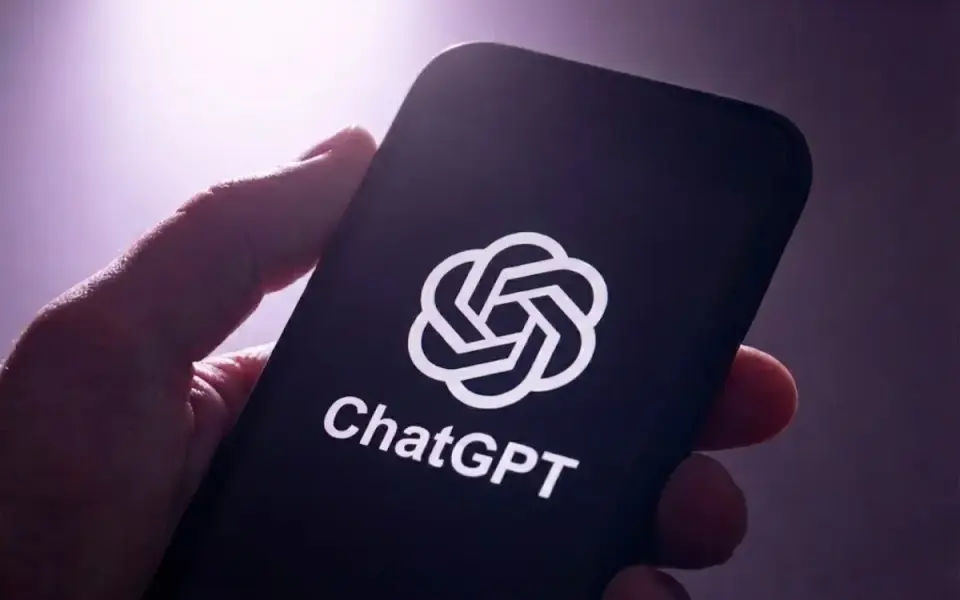 Mão segurando um celular com o logotipo do ChatGPT na tela em um ambiente escuro com luz roxa