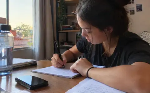 Estudante escrevendo uma redação à mão em folha pautada, à mesa de casa, com garrafa de água, caderno e celular ao lado e a luz do fim de tarde entrando pela janela
