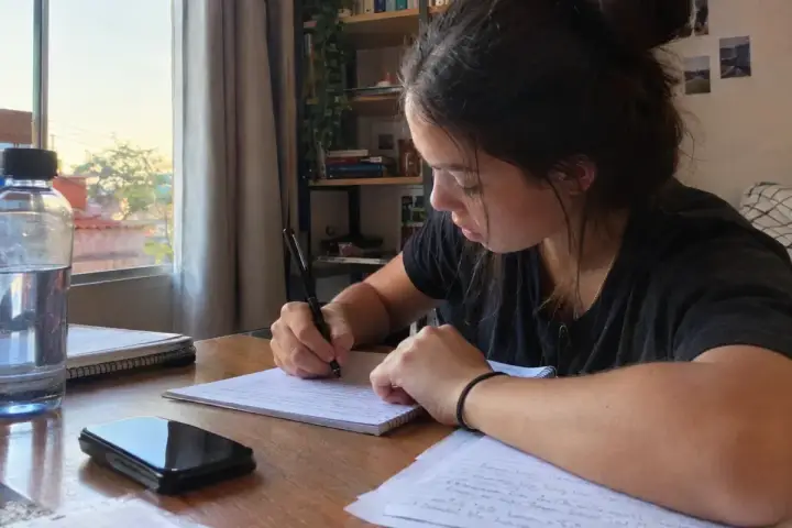 Estudante escrevendo uma redação à mão em folha pautada, à mesa de casa, com garrafa de água, caderno e celular ao lado e a luz do fim de tarde entrando pela janela