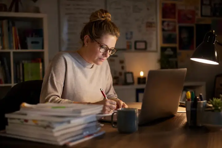 Professora corrigindo atividades à noite na mesa de casa, com notebook aberto, pilha de cadernos e uma caneca de café