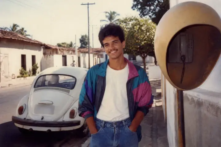 Homem brasileiro em foto no estilo anos 80, de jaqueta colorida e jeans, em uma rua de bairro com fusca branco e orelhão