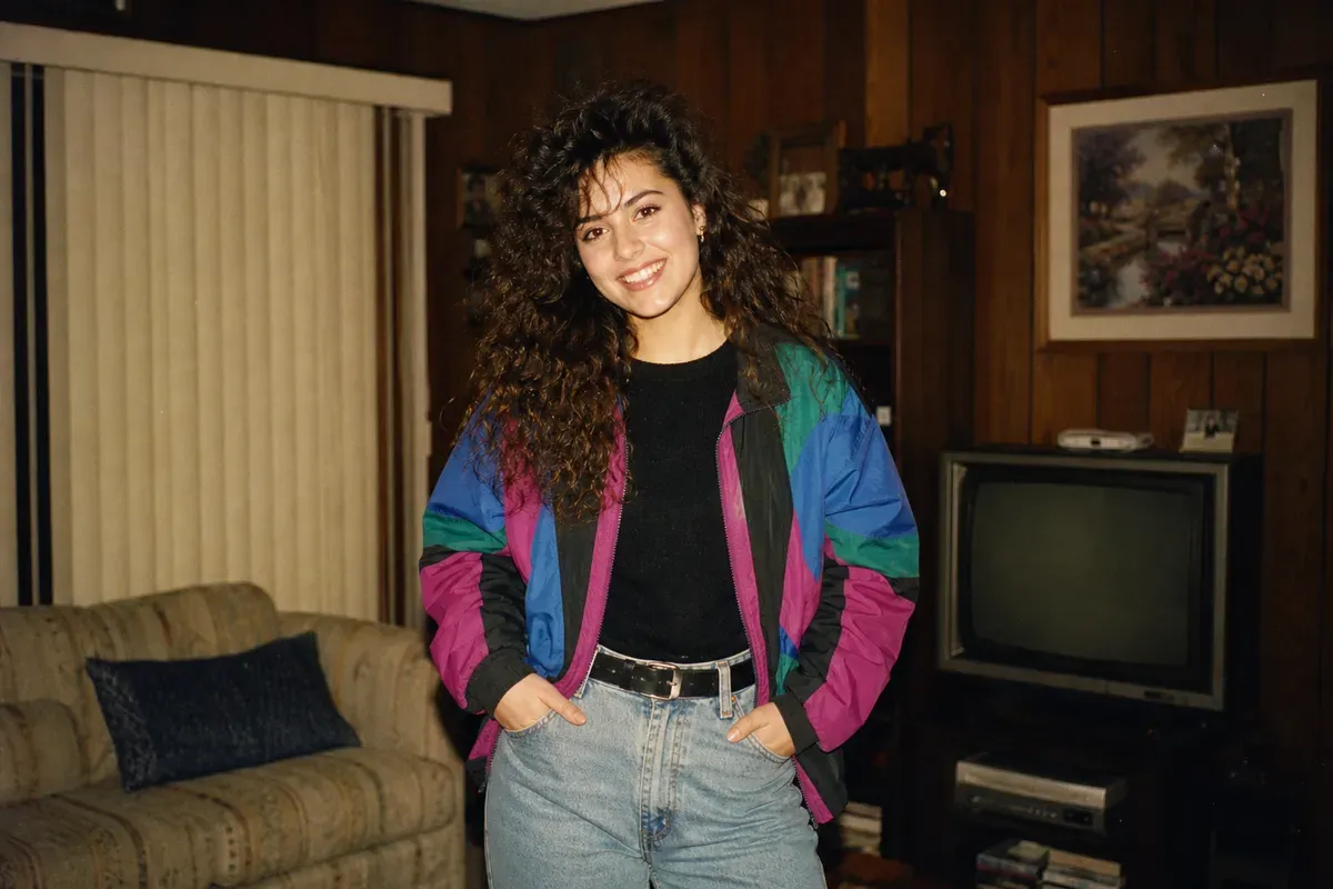 Retrato no estilo de foto de família dos anos 80, com jovem de cabelo cacheado volumoso, jaqueta colorida e jeans de cintura alta em uma sala com painéis de madeira e TV de tubo